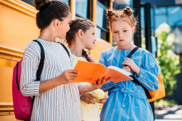 Group of little schoolgirls with notebook talking in front of school bus — Stock Photo