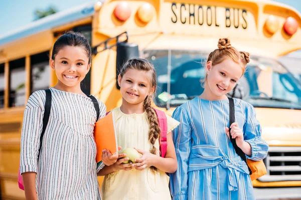 Счастливые маленькие школьницы смотрят в камеру перед школьным автобусом — стоковое фото