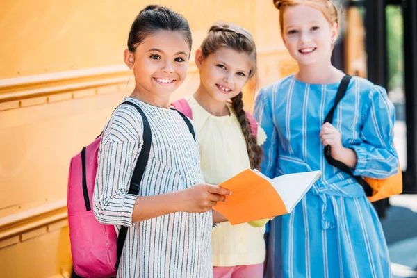 Adorable little schoolgirls with notebook looking at camera in front of school bus — Stock Photo