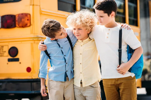 Fröhliche kleine Schüler, die sich vor dem Schulbus umarmen und unterhalten — Stockfoto