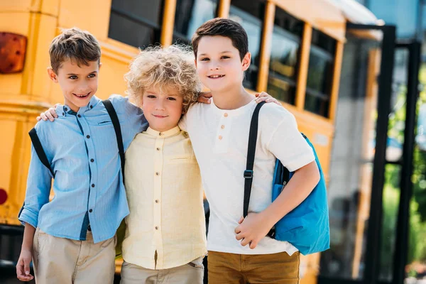 Adorable little schoolboys embracing in front of school bus and looking at camera — Stock Photo