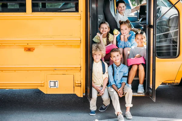 Gruppo di adorabili scolari seduti sulle scale dello scuolabus e guardando la macchina fotografica — Foto stock