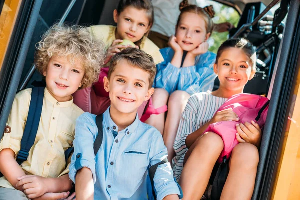 Grupo de escolares sonrientes sentados en las escaleras del autobús escolar y mirando a la cámara - foto de stock