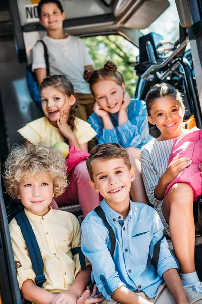 Grupo de alumnos adorables sentados en las escaleras del autobús escolar y mirando a la cámara - foto de stock