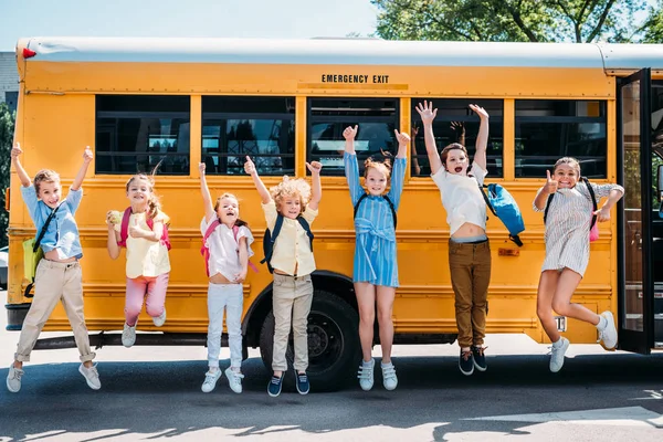 Grupo de alunos felizes pulando na frente do ônibus escolar e olhando para a câmera — Fotografia de Stock