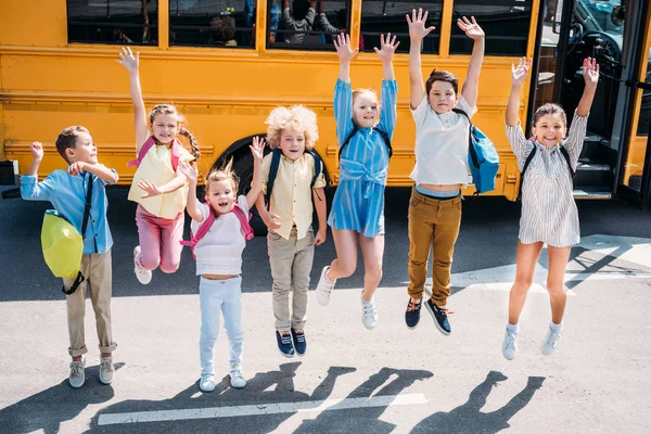 Grupo de escolares adorables saltando frente al autobús escolar y mirando a la cámara - foto de stock