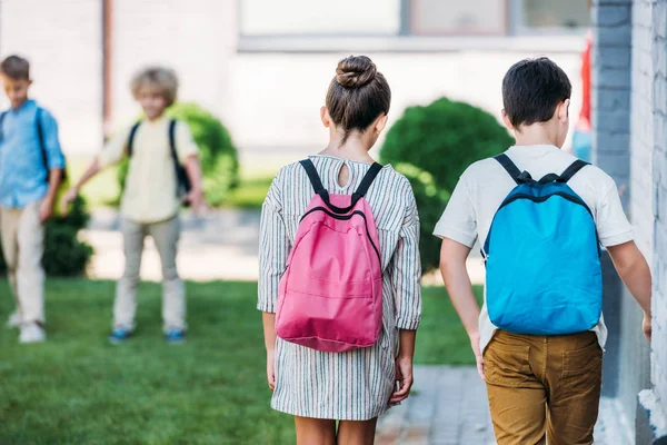 Vista trasera de los escolares con bolsitas caminando por el jardín escolar - foto de stock