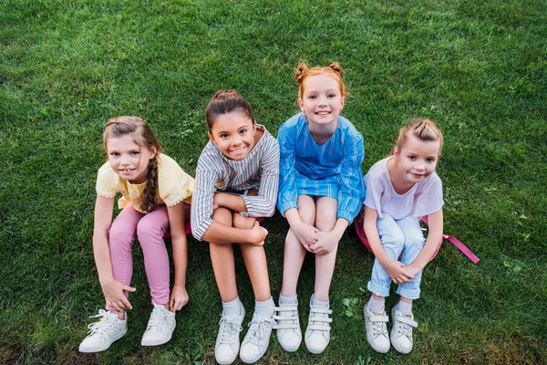 High angle view of group of happy schoolgirls sitting on green grass together and looking at camera — Stock Photo