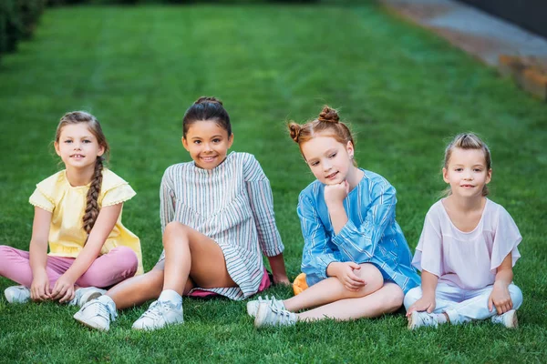 Група чарівних школярки сидять на зеленій траві разом і дивляться на камеру — стокове фото