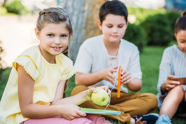 Adorable petite écolière avec pomme et livre assis sur l'herbe avec des camarades de classe — Photo de stock