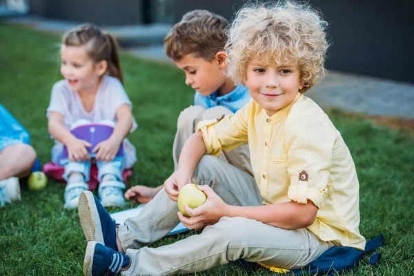 Adorable curly schoolboy with apple sitting on grass with classmates — Stock Photo