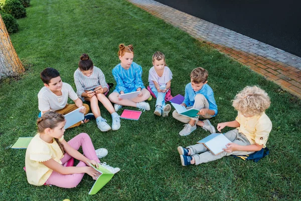 Высокий угол обзора группы учащихся, сидящих на траве с книгами и устройствами — стоковое фото