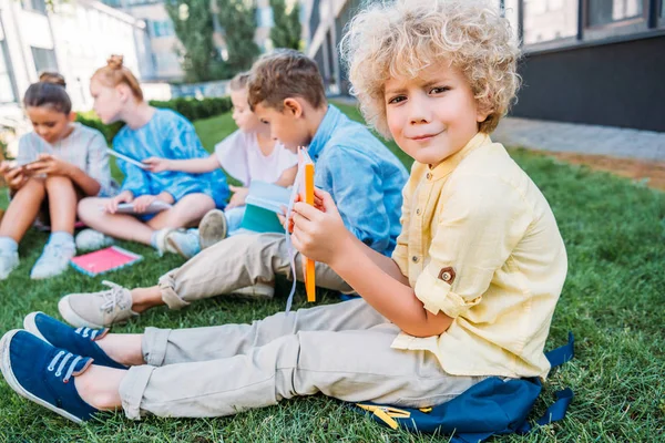 Confused curly schoolboy holding book while sitting on grass with classmates — Stock Photo