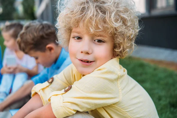 Close-up portrait of adorable curly schoolboy sitting on grass with classmates — Stock Photo