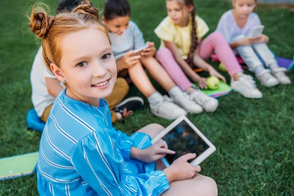 Sourire adorable écolière en utilisant la tablette tout en étant assis sur l'herbe avec des camarades de classe — Photo de stock