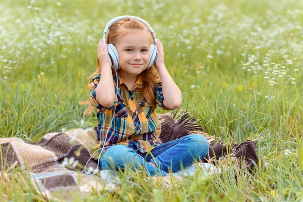 Lindo niño escuchando música en auriculares mientras está sentado en la manta en el campo con flores silvestres — Stock Photo