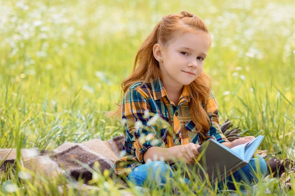 Retrato de adorable niño de pelo rojo con libro sentado en el campo de verano - foto de stock