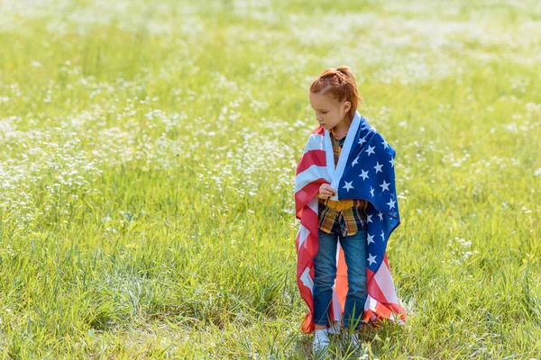 Criança de cabelo vermelho coberto de bandeira americana em pé no campo de verão — Fotografia de Stock