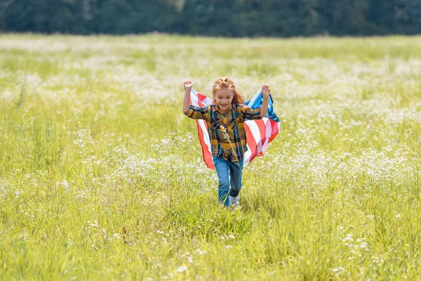 Niño alegre corriendo en el campo con bandera americana en las manos - foto de stock