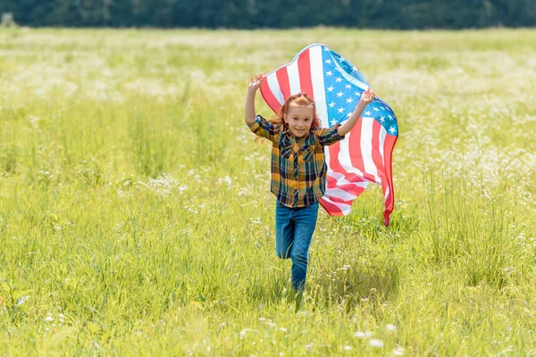 Niño alegre corriendo en el campo con bandera americana en las manos - foto de stock