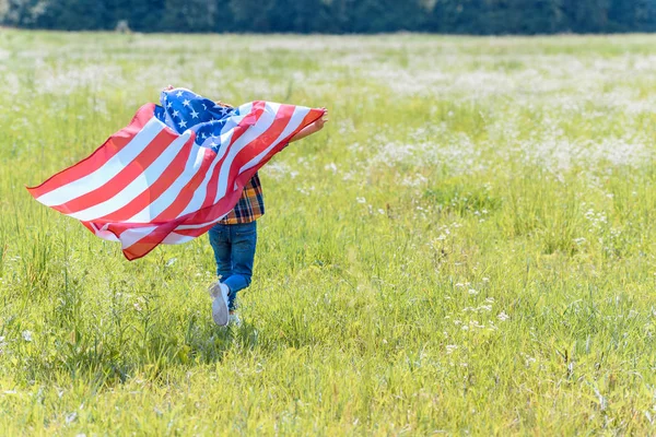 Visão traseira da criança correndo em campo com bandeira americana em mãos — Fotografia de Stock