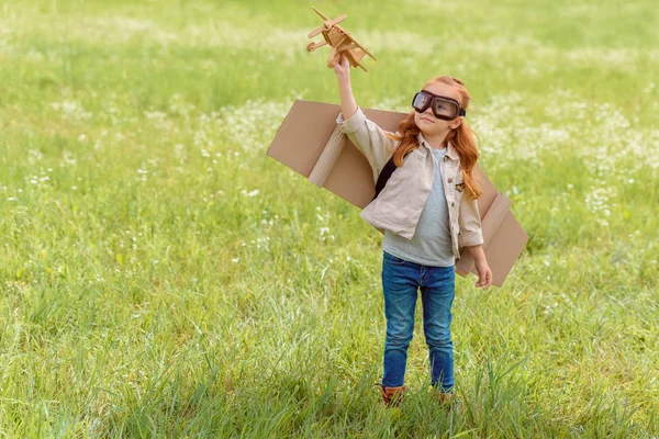 Porträt eines kleinen Kindes im Pilotenkostüm mit hölzernem Spielzeugflugzeug, das auf einer Wiese steht — Stockfoto