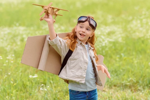 Портрет маленької дитини в пілотному костюмі з дерев'яною іграшковою площиною, що стоїть на лузі — стокове фото