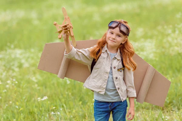 Porträt eines kleinen Kindes im Pilotenkostüm mit hölzernem Spielzeugflugzeug, das auf einer Wiese steht — Stockfoto