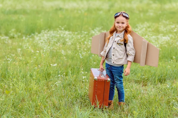 Adorable kid in pilot costume with retro suitcase standing in summer field — Stock Photo