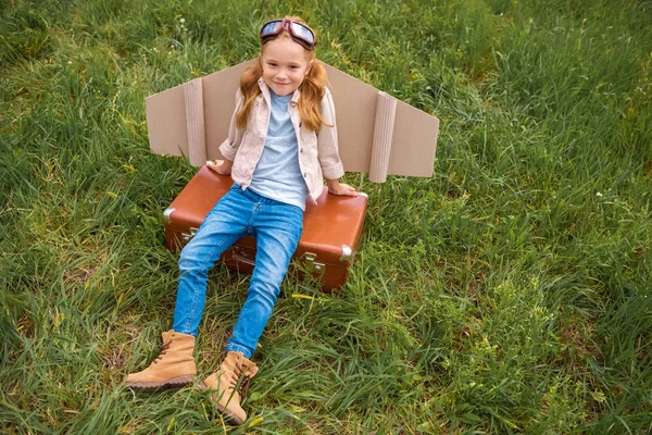 High angle view of smiling child with paper plane wings and protective eyeglasses resting on retro suitcase in meadow — Stock Photo