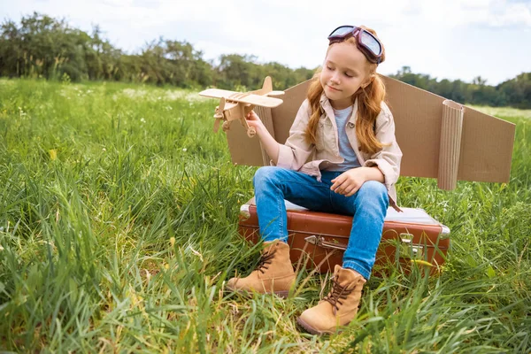 Kid in pilot costume with wooden toy plane in hand sitting on retro suitcase in field — Stock Photo