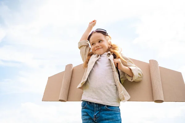 Vista basso angolo di bambino sorridente in costume da pilota con mano contro cielo nuvoloso blu — Foto stock