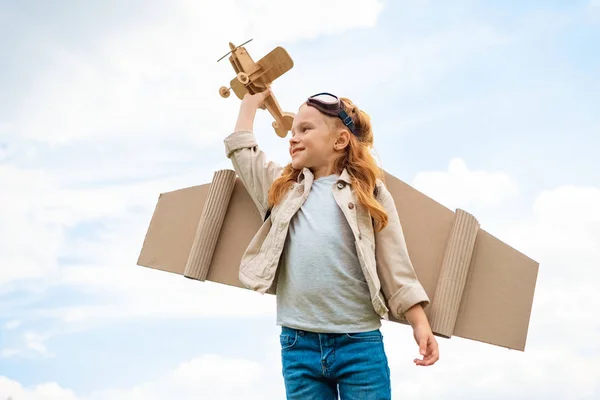 Vista de ángulo bajo del niño en traje de piloto sosteniendo avión de juguete de madera contra el cielo azul nublado - foto de stock