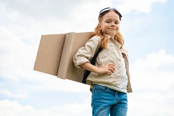 Vista de ángulo bajo del niño con alas planas de papel en la espalda y gafas protectoras en la cabeza contra el cielo azul nublado - foto de stock