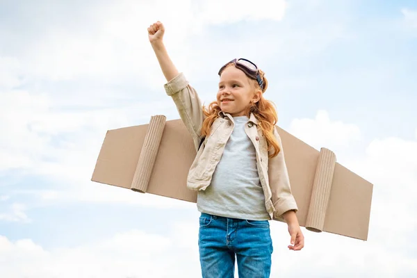 Retrato de niño con alas planas de papel en la espalda y gafas protectoras en la cabeza de pie con el brazo extendido contra el cielo azul nublado - foto de stock