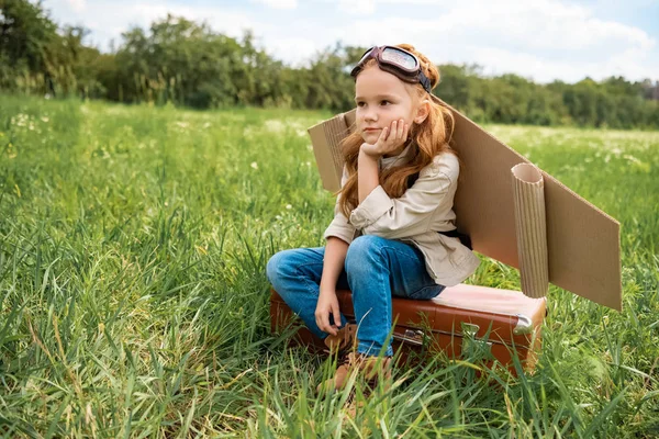 Задумчивый ребенок в костюме пилота сидит на ретро-чемодане в летнем поле — стоковое фото