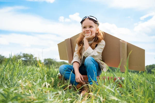 Adorable kid in pilot costume sitting on retro suitcase in summer field with blue cloudy sky on background — Stock Photo