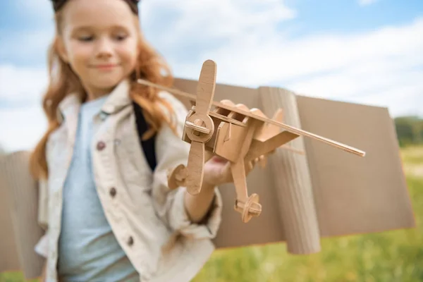 Selektiver Fokus eines Rothaarkindes im Pilotenkostüm, das Holzflugzeug gegen den blauen Himmel hält — Stockfoto