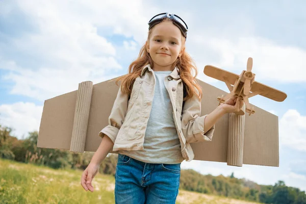 Porträt eines lächelnden Rothaarkindes im Pilotenkostüm, das ein Holzflugzeug im Sommerfeld hält — Stockfoto