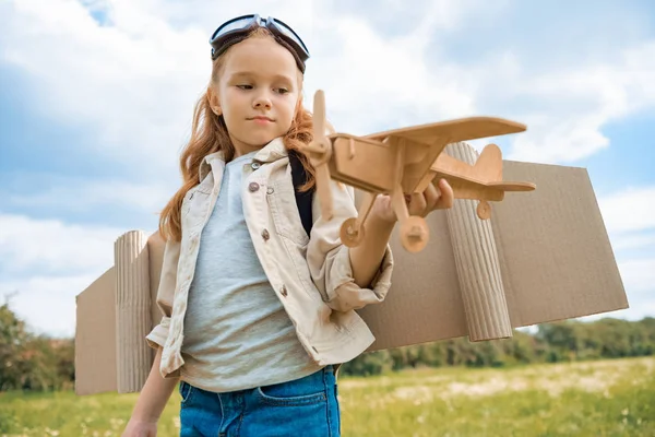 Ritratto di bambino con i capelli rossi in costume da pilota che tiene un aereo di legno nel campo estivo — Foto stock