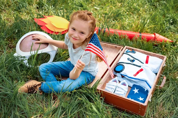 Улыбающийся ребенок с американским флагштоком в руке сидит рядом с чемоданом в костюме космонавта и ракетой на зеленой траве — стоковое фото