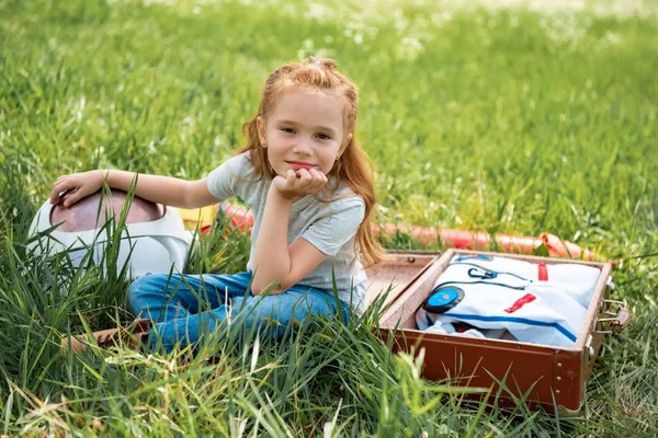 Smiling red hair kid sitting near suitcase with cosmonaut costume and rocket on green grass — Stock Photo