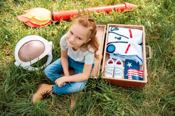 High angle view of smiling red hair kid sitting near suitcase with cosmonaut costume and rocket on green grass — Stock Photo