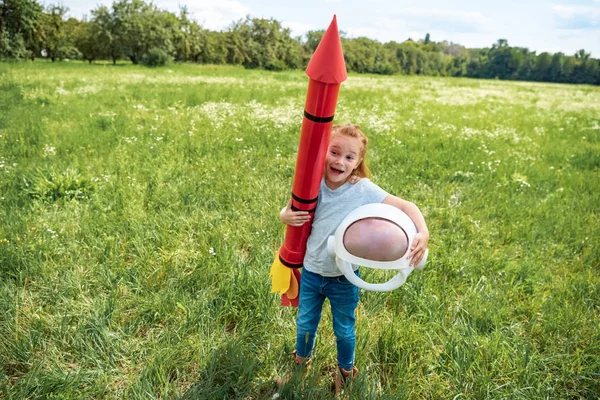 Cabellera roja excitada con cohete y casco astronauta de pie en el campo de verano - foto de stock
