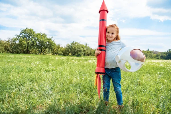 Lächelndes Rothaarkind mit Rakete und Astronautenhelm steht im Sommerfeld — Stockfoto