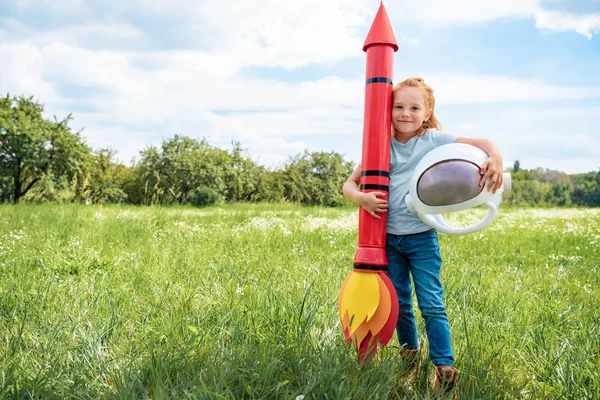 Sonriente niño de pelo rojo con cohete y casco astronauta de pie en el campo de verano - foto de stock