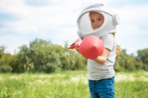 Portrait of kid in cosmonaut helmet with rocket in hands standing in field — Stock Photo