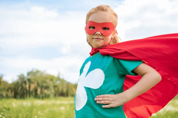 Portrait of little child in red superhero costume standing akimbo in summer meadow — Stock Photo