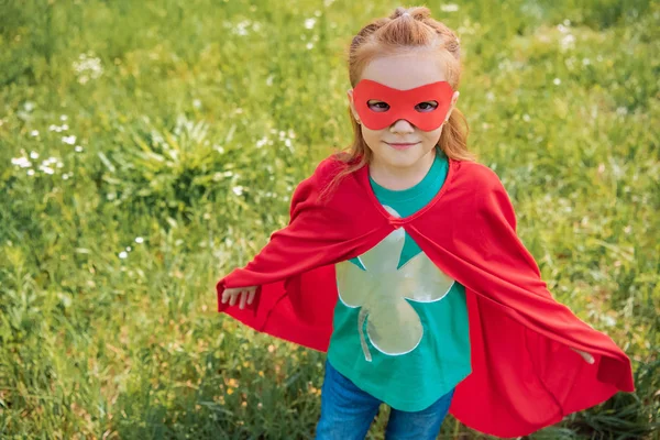 Cute child in red superhero mask and cape standing in summer field — Stock Photo