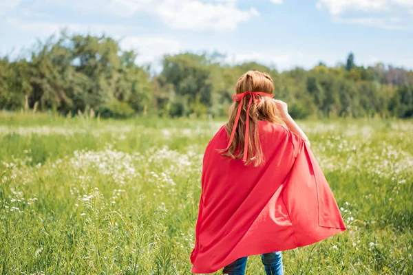 Visão traseira da criança em capa de super-herói vermelho e máscara em pé no campo de verão — Fotografia de Stock